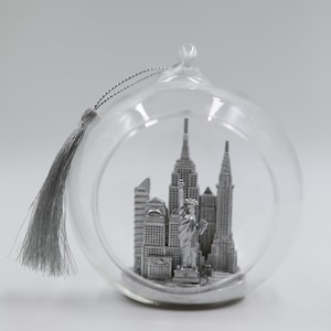 Puede incluir: Una bola de Navidad de cristal transparente con una borla y una cuerda plateadas. Dentro de la bola hay una miniatura plateada del horizonte de la ciudad de Nueva York, incluyendo la Estatua de la Libertad y el Empire State Building.