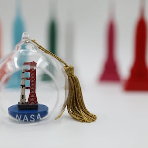 Può includere: Un ornamento di vetro con un fiocco dorato presenta un modello in miniatura di un lancio di razzi con la scritta "NASA" sulla base.