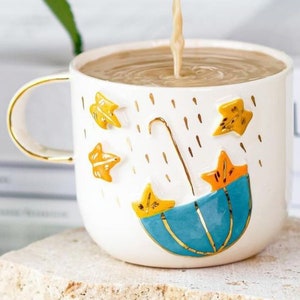 May include: A white ceramic mug with a gold rim and handle. The mug has a blue umbrella and yellow stars design.  The mug is filled with a light brown liquid.