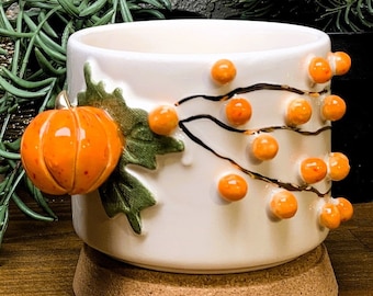 May include: White ceramic mug with a 3D pumpkin and orange berries design. The mug has a cork base.