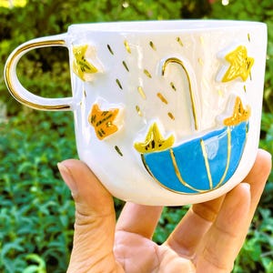 May include: White ceramic mug with a blue and gold umbrella design, gold stars, and gold raindrops. The mug has a gold handle.