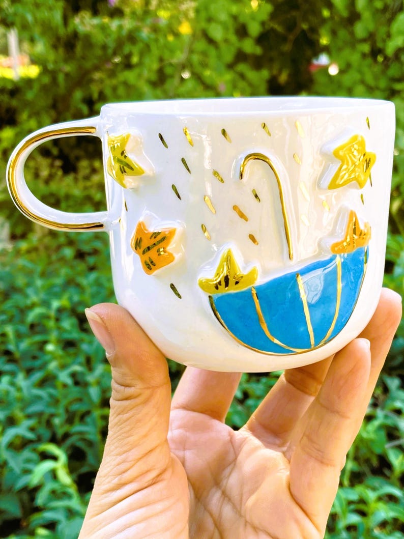 May include: White ceramic mug with a blue and gold umbrella design, gold stars, and gold raindrops. The mug has a gold handle.