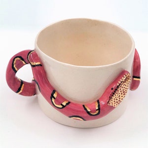 Könnte beinhalten: Cremefarbene Keramiktasse mit einer roten Schlange. Die Schlange hat schwarze und goldene Akzente, der Kopf bildet den Griff. Ein einzigartiges, handgefertigtes Stück.