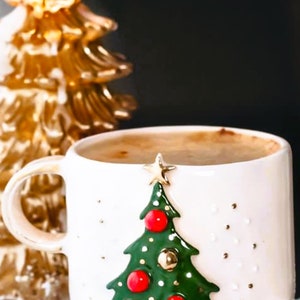 May include: White ceramic mug with a green Christmas tree design and red and gold ornaments. The mug is filled with a brown liquid and is sitting on a white surface next to a gold Christmas tree candle.