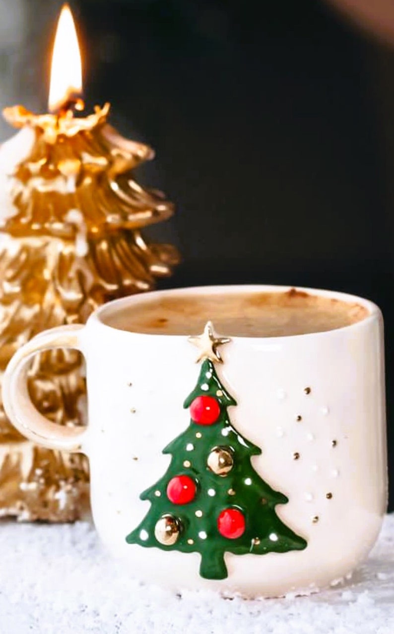 May include: White ceramic mug with a green Christmas tree design and red and gold ornaments. The mug is filled with a brown liquid and is sitting on a white surface next to a gold Christmas tree candle.