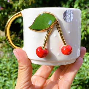 Pu&ograve; includere: Tazza in ceramica bianca con manico dorato e design a ciliegie 3D. La ciliegia ha una foglia verde e steli dorati. La tazza ha un motivo a pois bianchi.