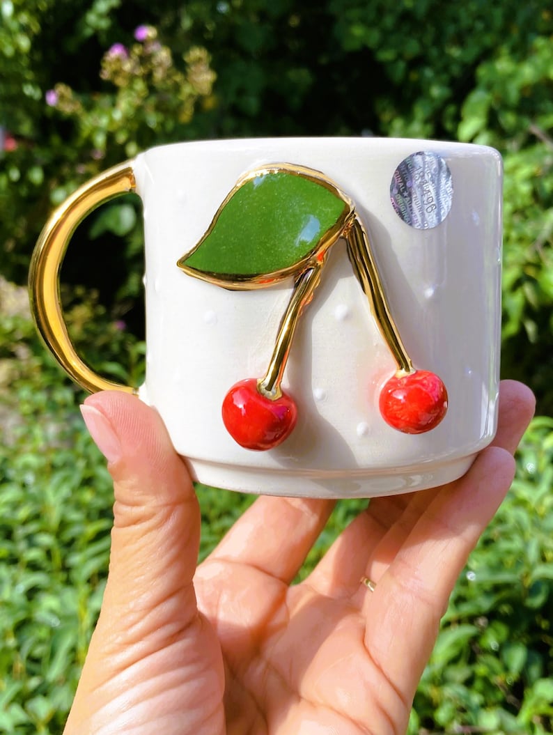 Pu&ograve; includere: Tazza in ceramica bianca con manico dorato e design a ciliegie 3D. La ciliegia ha una foglia verde e steli dorati. La tazza ha un motivo a pois bianchi.