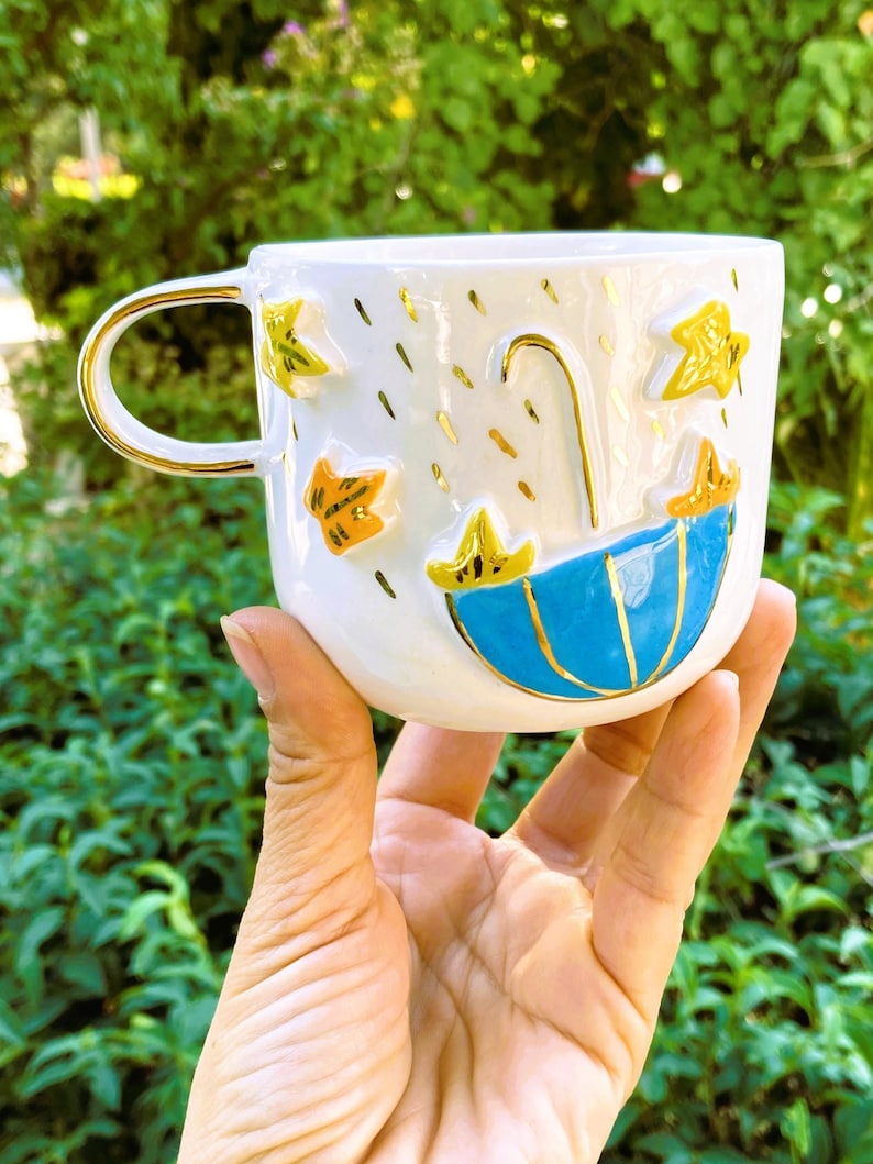 May include: White ceramic mug with a blue and gold umbrella design. The mug has a gold handle and is decorated with yellow and orange stars and raindrops.