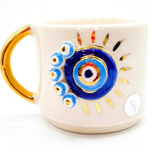 K&ouml;nnte beinhalten: Wei&szlig;e Keramiktasse mit goldenem Henkel und einem blauen und goldenen b&ouml;sen Auge. Die Tasse hat ein goldenes Sonnenstrahlen-Design und das Wort "Ceramika" ist auf dem Boden geschrieben.