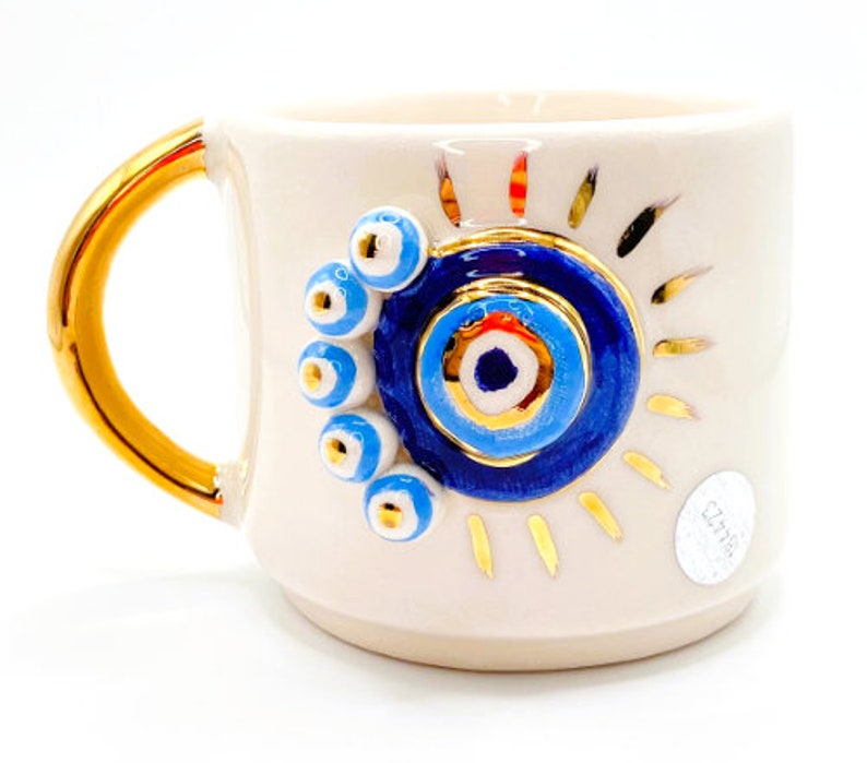 K&ouml;nnte beinhalten: Wei&szlig;e Keramiktasse mit goldenem Henkel und einem blauen und goldenen b&ouml;sen Auge. Die Tasse hat ein goldenes Sonnenstrahlen-Design und das Wort "Ceramika" ist auf dem Boden geschrieben.