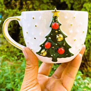 K&ouml;nnte beinhalten: Wei&szlig;e Keramik-Weihnachtstasse mit goldenem Henkel und Rand. Die Tasse hat ein gr&uuml;nes Weihnachtsbaumdesign mit roten und goldenen Ornamenten. Goldene Tupfen sind &uuml;ber die Oberfl&auml;che verstreut. Ein goldener Stern kr&ouml;nt den Baum.
