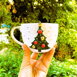 May include: White ceramic mug with a green Christmas tree design. The tree has red ornaments and gold bells. The mug has a gold handle.