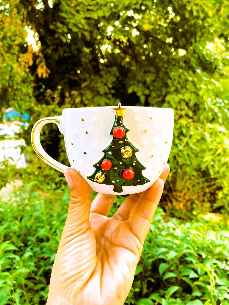 May include: White ceramic mug with a green Christmas tree design. The tree has red ornaments and gold bells. The mug has a gold handle.