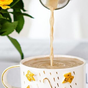 May include: A white ceramic mug with a gold rim and handle. The mug has a blue and yellow umbrella design with gold accents and a pattern of yellow leaves and gold raindrops. The mug is filled with a light brown liquid being poured from a glass pitcher.