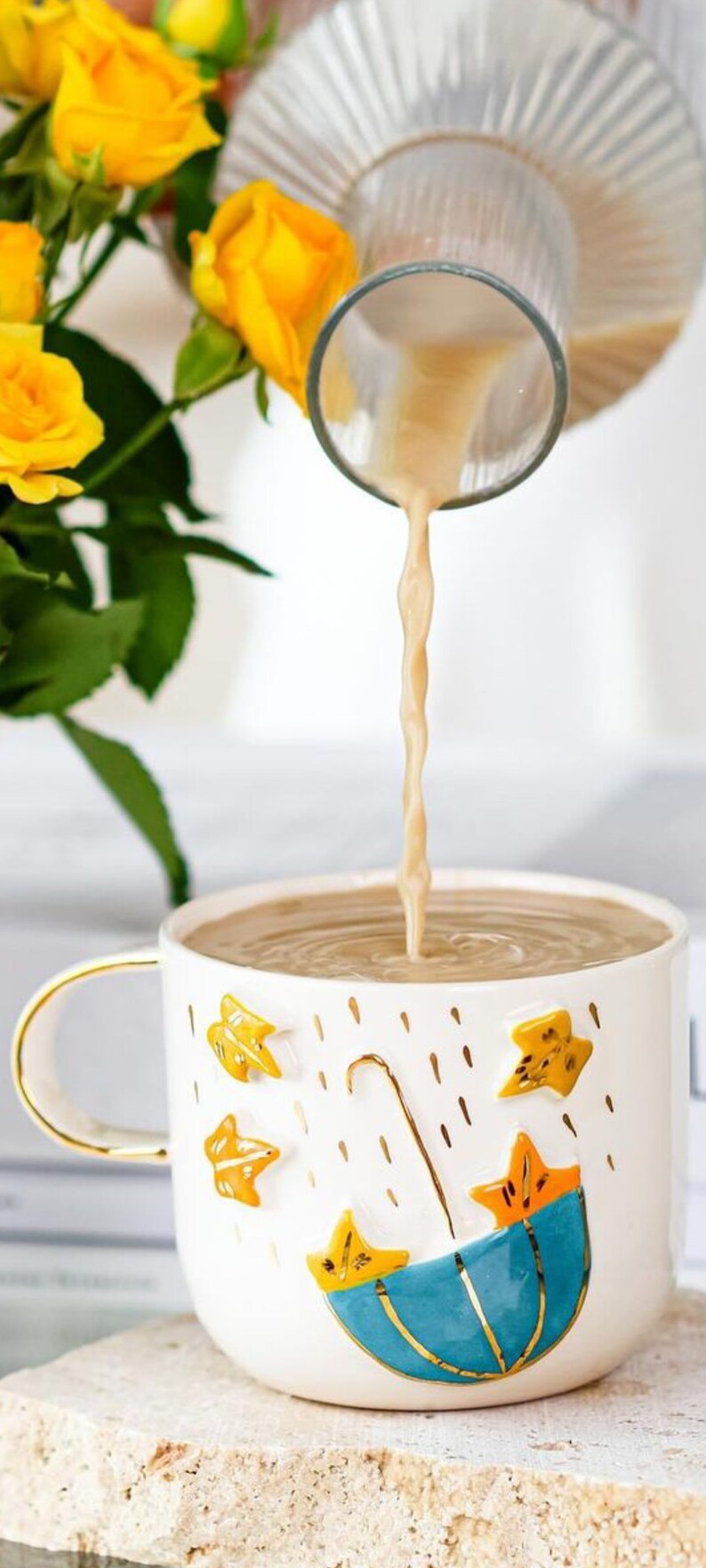 May include: A white ceramic mug with a gold rim and handle. The mug has a blue and yellow umbrella design with gold accents and a pattern of yellow leaves and gold raindrops. The mug is filled with a light brown liquid being poured from a glass pitcher.
