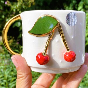 Pu&ograve; includere: Tazza in ceramica bianca con manico dorato e due ciliegie rosse con foglie verdi. La tazza ha un motivo a pois bianchi.