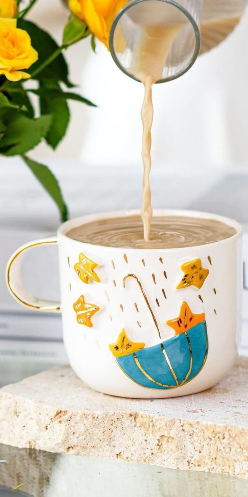 May include: A white ceramic mug with a gold rim and handle. The mug has a blue umbrella and yellow stars design.  The mug is filled with a light brown liquid.