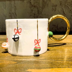 K&ouml;nnte beinhalten: Wei&szlig;e Keramiktasse mit goldenem Henkel, verziert mit h&auml;ngenden Kugeln und roten Schleifen. Die Kugeln sind in verschiedenen Farben und Mustern gehalten. Eine festliche Kaffeetasse zum Thema Feiertage.