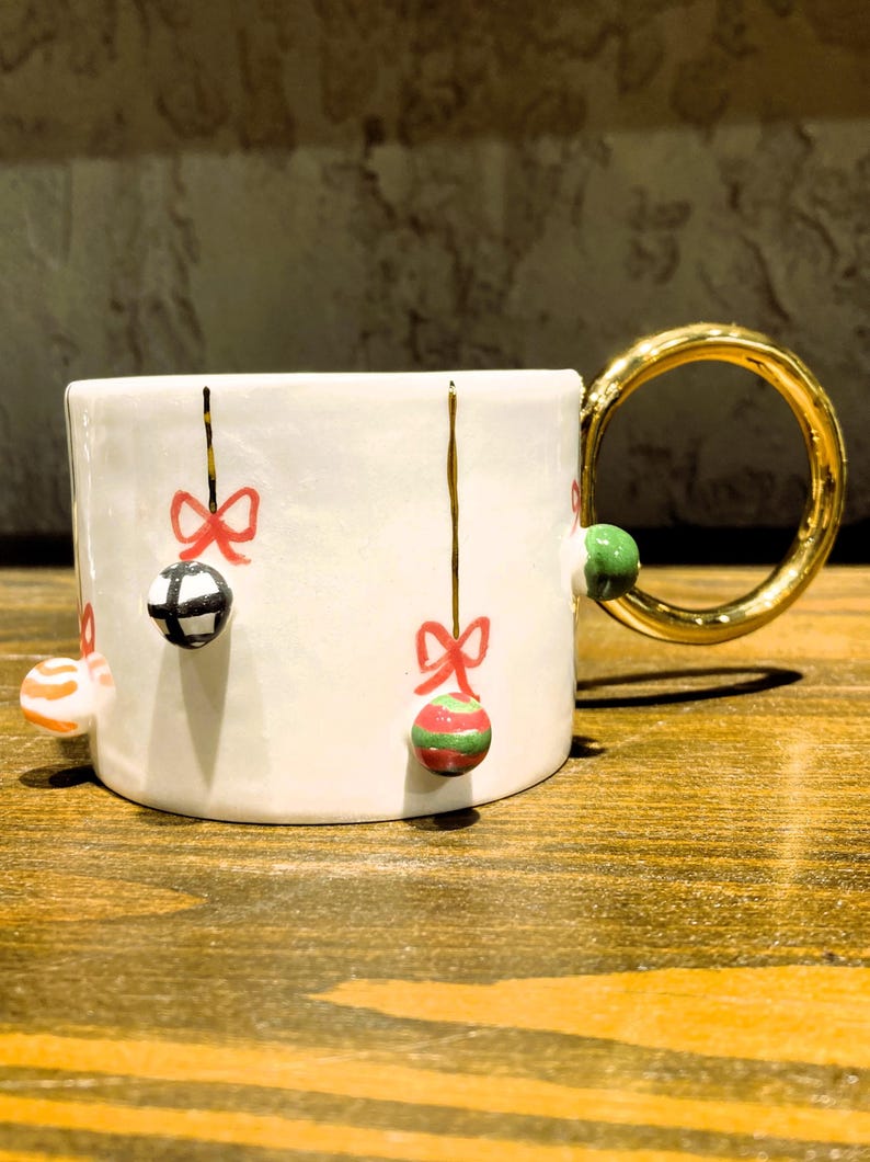 K&ouml;nnte beinhalten: Wei&szlig;e Keramiktasse mit goldenem Henkel, verziert mit h&auml;ngenden Kugeln und roten Schleifen. Die Kugeln sind in verschiedenen Farben und Mustern gehalten. Eine festliche Kaffeetasse zum Thema Feiertage.