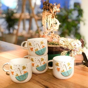 May include: Four white ceramic mugs with a fall theme. Each mug features a blue umbrella with a gold handle, yellow and orange leaves, and gold raindrops.