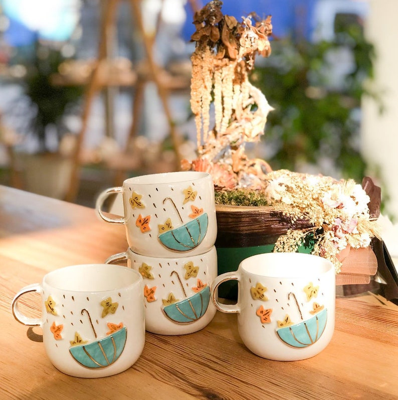 May include: Four white ceramic mugs with a fall theme. Each mug features a blue umbrella with a gold handle, yellow and orange leaves, and gold raindrops.