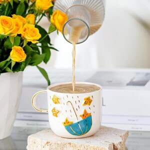 May include: A white ceramic mug with a blue umbrella and yellow stars design.  The mug is filled with a light brown liquid being poured from a glass pitcher.