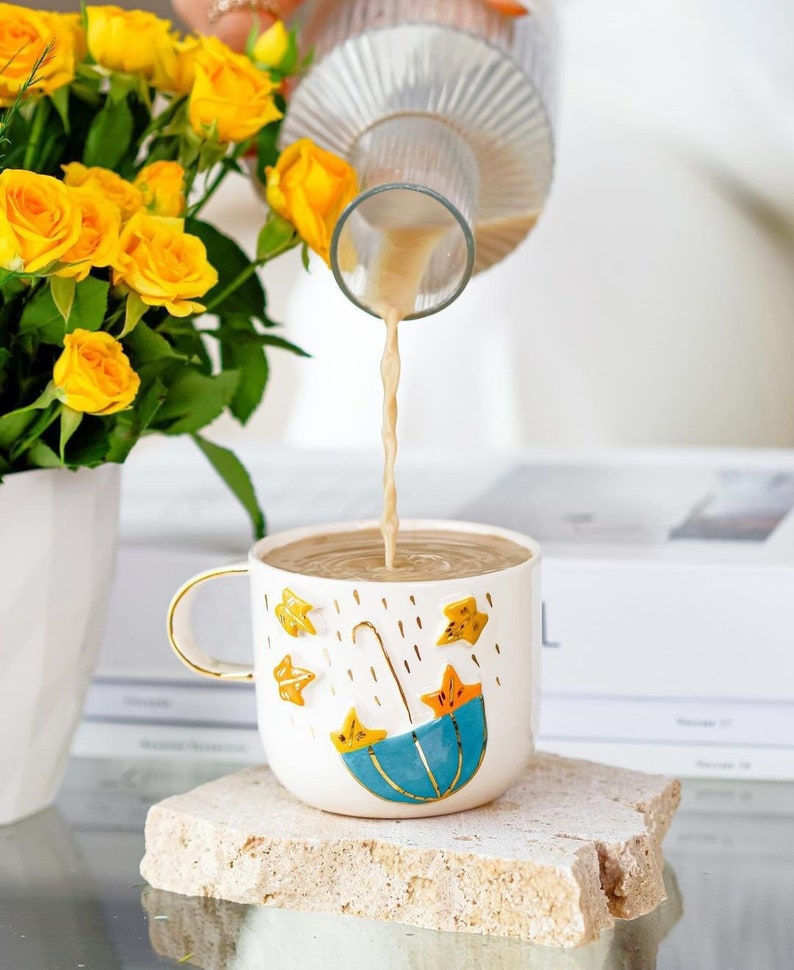 May include: A white ceramic mug with a blue umbrella and yellow stars design.  The mug is filled with a light brown liquid being poured from a glass pitcher.