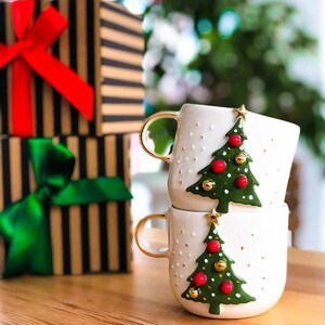 May include: Two white ceramic mugs with gold handles and green Christmas tree designs. The trees have red and gold ornaments. The mugs are stacked on top of each other.