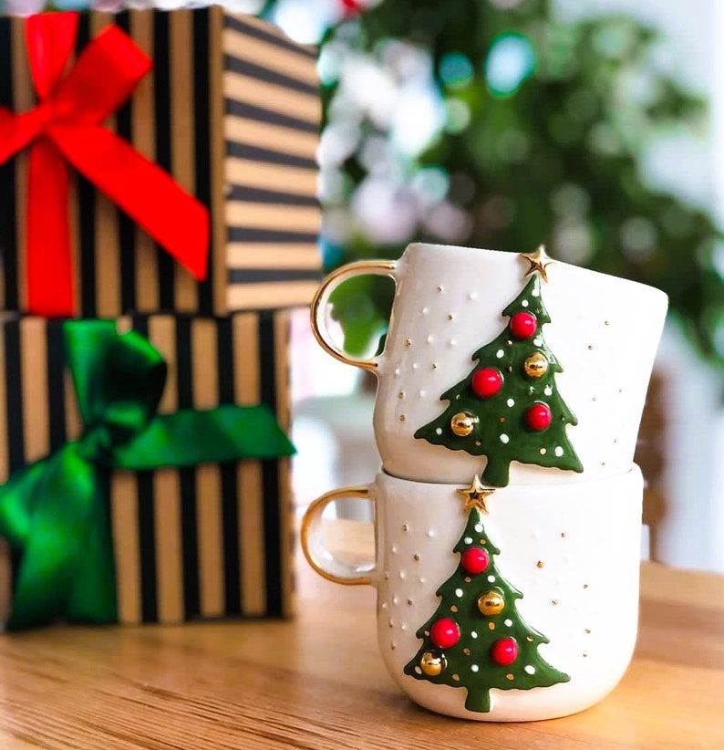 May include: Two white ceramic mugs with gold handles and green Christmas tree designs. The trees have red and gold ornaments. The mugs are stacked on top of each other.