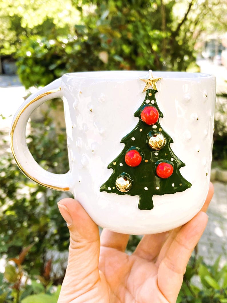 K&ouml;nnte beinhalten: Wei&szlig;e Keramiktasse mit goldenem Henkel und Rand, verziert mit einem gr&uuml;nen Weihnachtsbaum-Design. Der Baum ist mit roten und goldenen Kugeln geschm&uuml;ckt und mit einem goldenen Stern gekr&ouml;nt. Die Tasse hat eine strukturierte Oberfl&auml;che mit goldenen Sprenkeln.