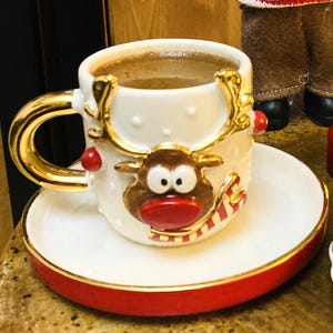 K&ouml;nnte beinhalten: Ein wei&szlig;es Keramik-Kaffeebecher- und Untertassen-Set mit einem festlichen Rentier-Design. Die Tasse hat einen goldenen Griff und Rand, und die Untertasse hat einen roten Rand. Die Tasse ist mit Kaffee gef&uuml;llt.