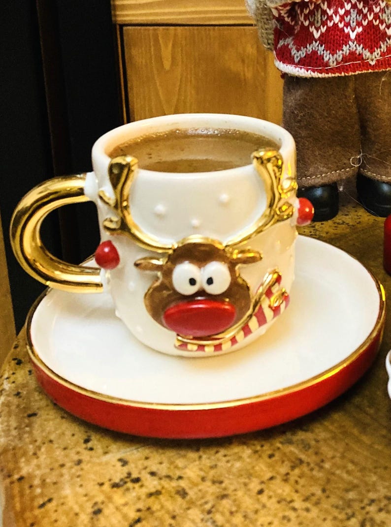 K&ouml;nnte beinhalten: Ein wei&szlig;es Keramik-Kaffeebecher- und Untertassen-Set mit einem festlichen Rentier-Design. Die Tasse hat einen goldenen Griff und Rand, und die Untertasse hat einen roten Rand. Die Tasse ist mit Kaffee gef&uuml;llt.