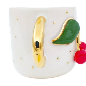 Pu&ograve; includere: Tazza in ceramica bianca con manico dorato e un design a ciliegie. Le ciliegie sono rosse con foglie verdi e sono dipinte sul lato della tazza.