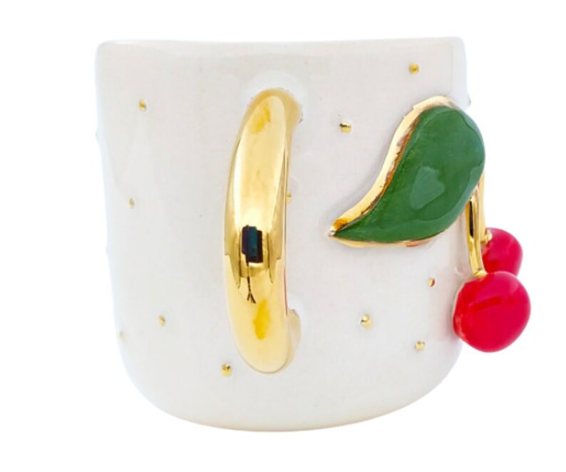 Pu&ograve; includere: Tazza in ceramica bianca con manico dorato e un design a ciliegie. Le ciliegie sono rosse con foglie verdi e sono dipinte sul lato della tazza.
