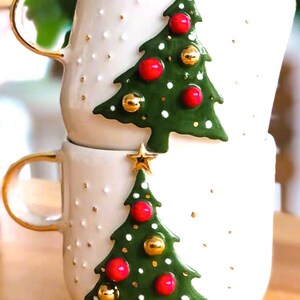 May include: Two white ceramic mugs with gold handles. Each mug features a 3D green Christmas tree with red and gold ornaments. The trees are decorated with white dots.