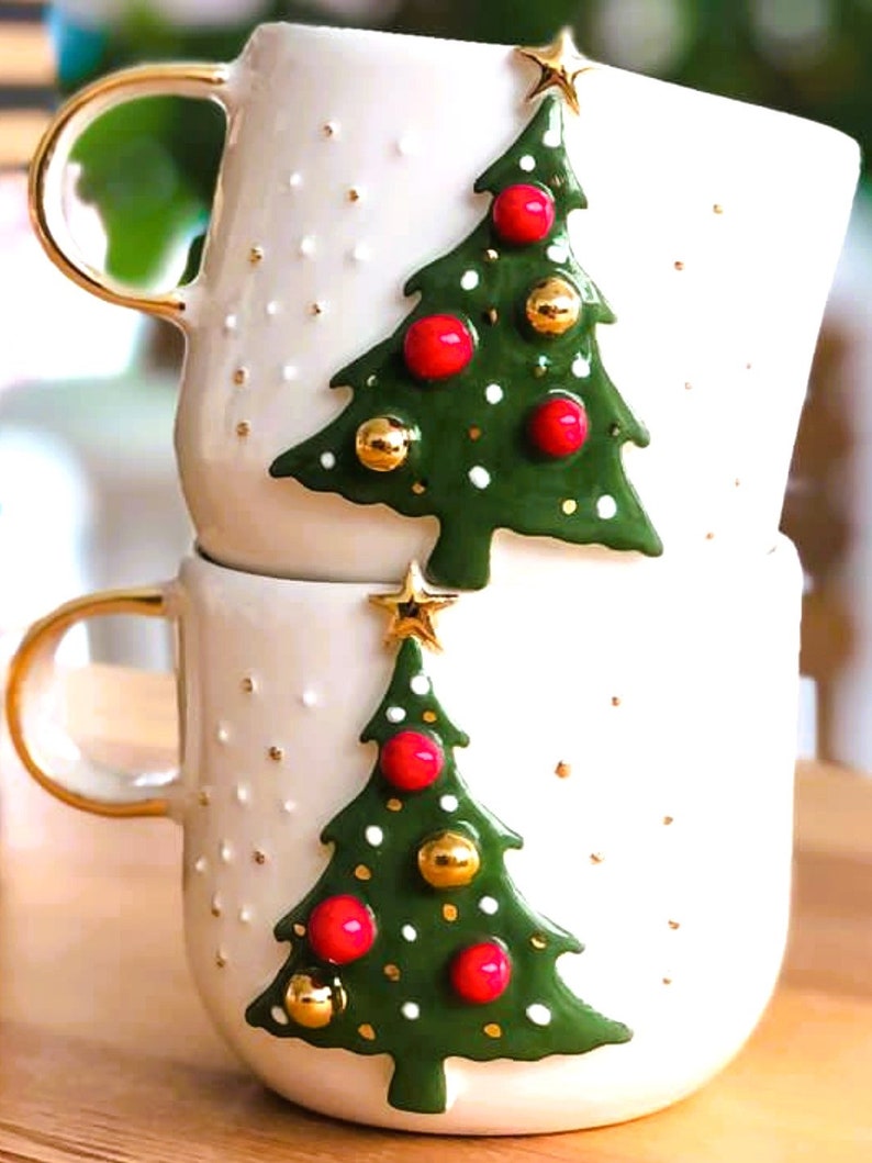 May include: Two white ceramic mugs with gold handles. Each mug features a 3D green Christmas tree with red and gold ornaments. The trees are decorated with white dots.
