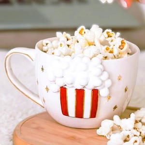 K&ouml;nnte beinhalten: Eine wei&szlig;e Keramik-Tasse gef&uuml;llt mit Popcorn, mit einem rot-wei&szlig;en Popcorn-Box-Design. Die Tasse hat einen goldenen Henkel und goldene Sterne. Popcorn quillt auf ein Holztablett.