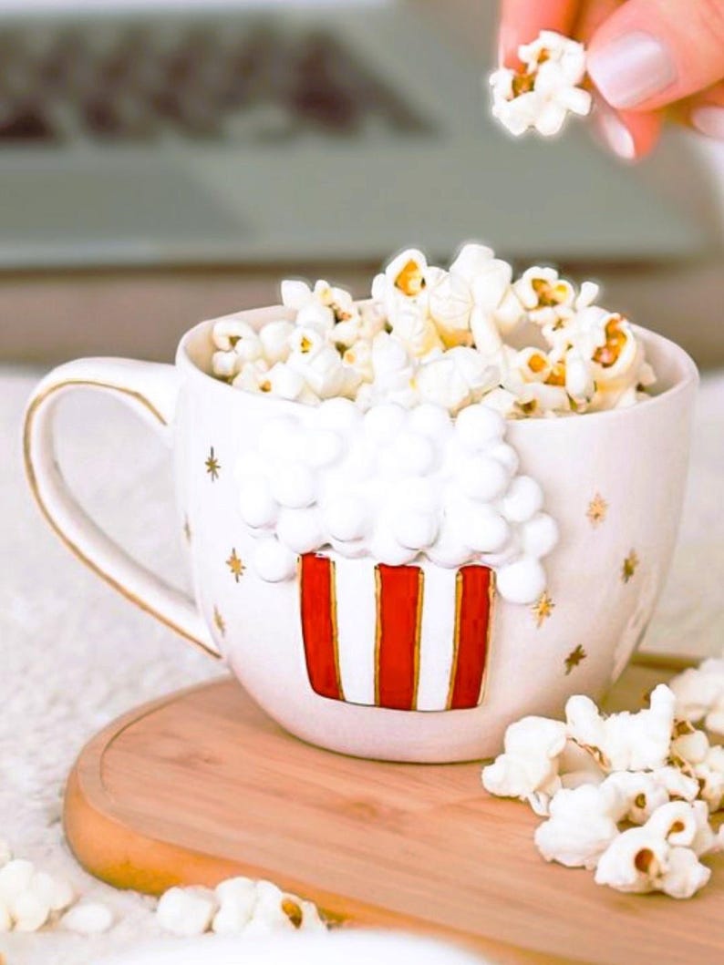K&ouml;nnte beinhalten: Eine wei&szlig;e Keramik-Tasse gef&uuml;llt mit Popcorn, mit einem rot-wei&szlig;en Popcorn-Box-Design. Die Tasse hat einen goldenen Henkel und goldene Sterne. Popcorn quillt auf ein Holztablett.