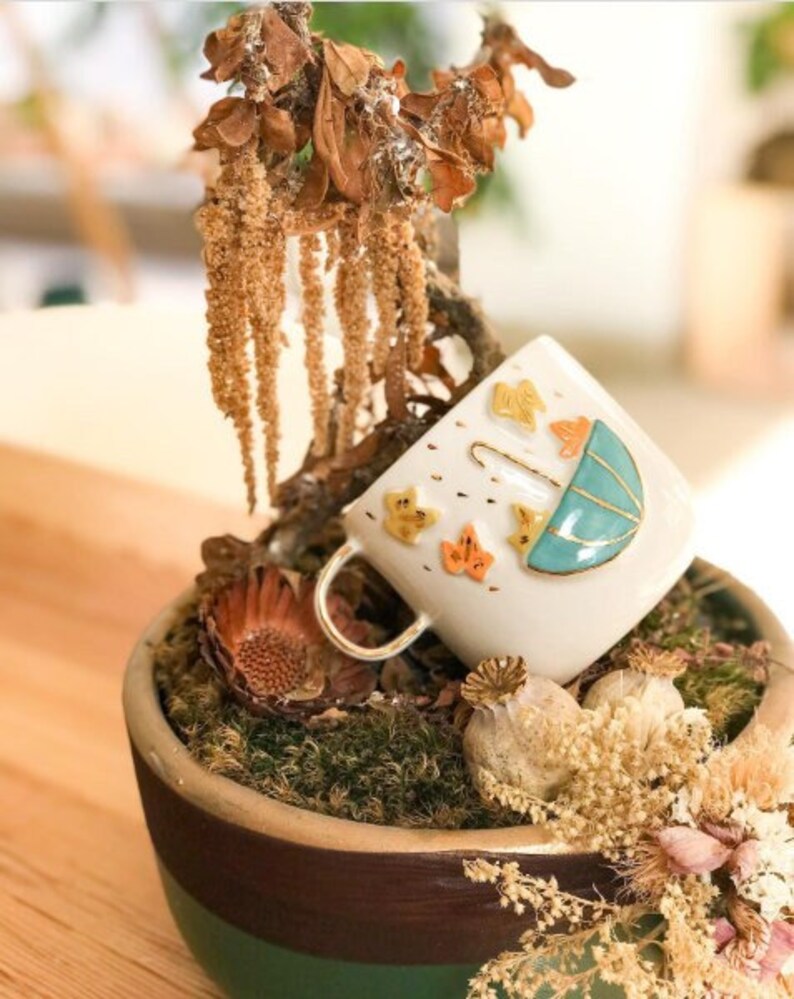 May include: A white ceramic mug with a teal blue umbrella and yellow and orange leaves painted on it. The mug is sitting in a brown and green planter with moss and dried flowers.