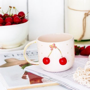 May include: A pink ceramic mug with a gold rim and handle. The mug has a white polka dot pattern and a gold cherry design with a pink bow.