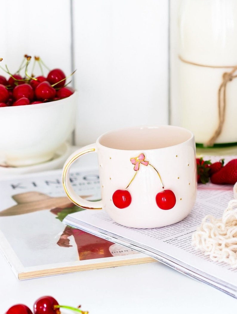 May include: A pink ceramic mug with a gold rim and handle. The mug has a white polka dot pattern and a gold cherry design with a pink bow.
