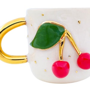 Pu&ograve; includere: Tazza in ceramica bianca con manico dorato e un design a ciliegie. Le ciliegie sono rosse con foglie verdi e steli dorati. La tazza ha uno sfondo bianco con macchie dorate.