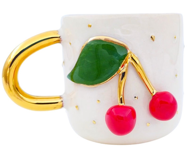 Pu&ograve; includere: Tazza in ceramica bianca con manico dorato e un design a ciliegie. Le ciliegie sono rosse con foglie verdi e steli dorati. La tazza ha uno sfondo bianco con macchie dorate.