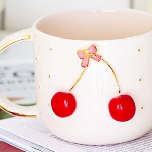May include: White ceramic mug with a gold rim and handle. The mug features a 3D design of two red cherries with a pink bow on top.