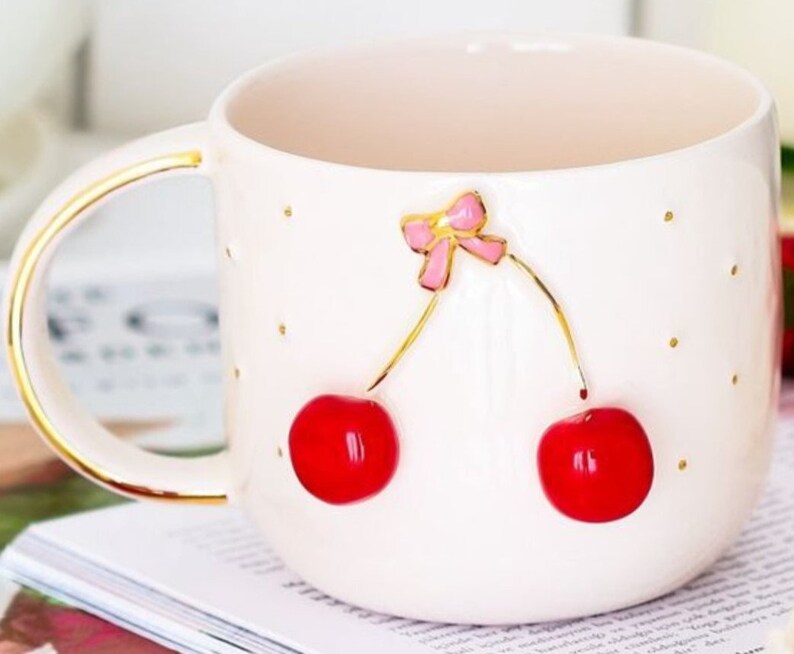 May include: White ceramic mug with a gold rim and handle. The mug features a 3D design of two red cherries with a pink bow on top.