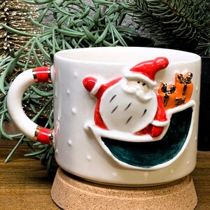 K&ouml;nnte beinhalten: Wei&szlig;e Keramik-Weihnachtstasse mit einem 3D-Santa-Claus-Design mit roten und wei&szlig;en Akzenten. Die Tasse hat ein gr&uuml;nes Innere und ist mit kleinen wei&szlig;en Punkten verziert. Der Griff hat rote und goldene Details. Die Tasse steht auf einem Korkuntersetzer.