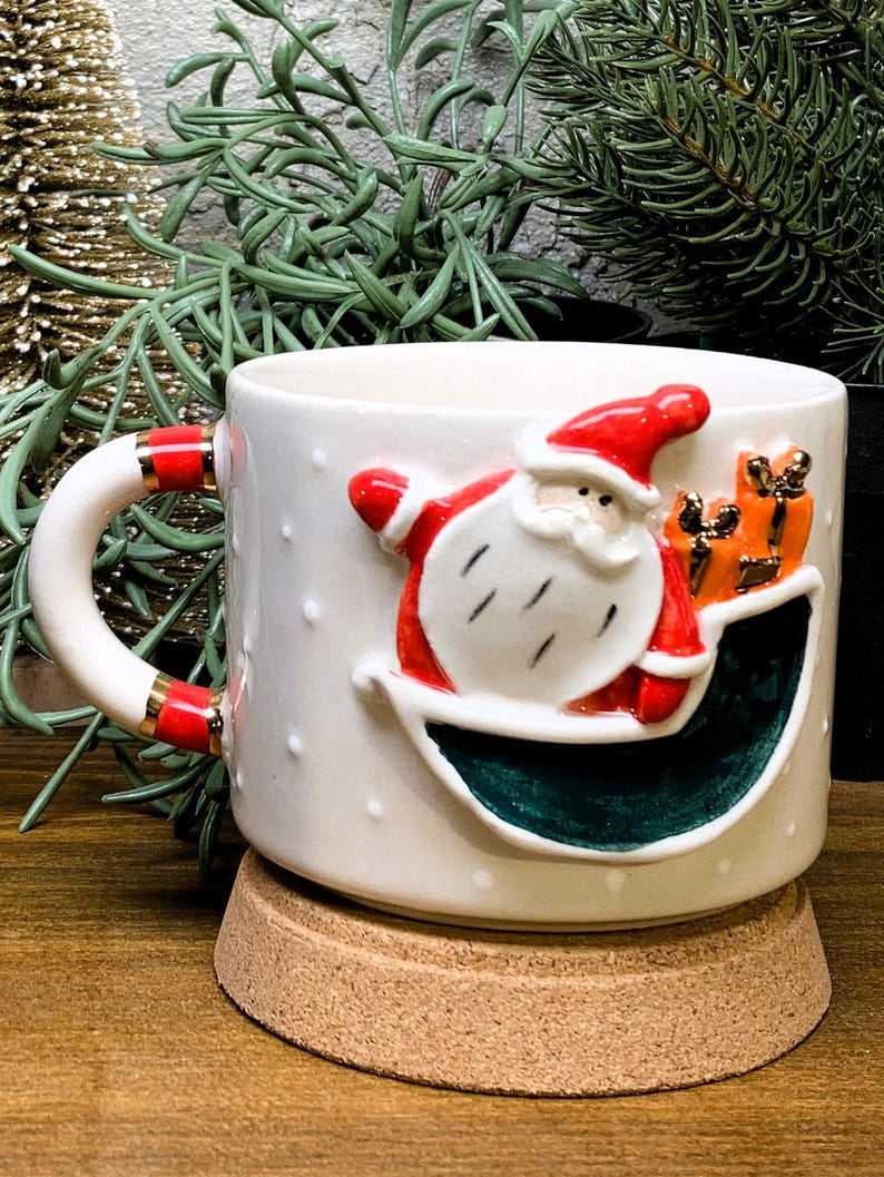 K&ouml;nnte beinhalten: Wei&szlig;e Keramik-Weihnachtstasse mit einem 3D-Santa-Claus-Design mit roten und wei&szlig;en Akzenten. Die Tasse hat ein gr&uuml;nes Innere und ist mit kleinen wei&szlig;en Punkten verziert. Der Griff hat rote und goldene Details. Die Tasse steht auf einem Korkuntersetzer.