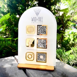 May include: A white and gold acrylic display stand with the text "WILD & FREE ARTISTRY". The stand features gold icons for a globe, Facebook, and Instagram, each paired with a QR code. The stand is set on a wooden base.
