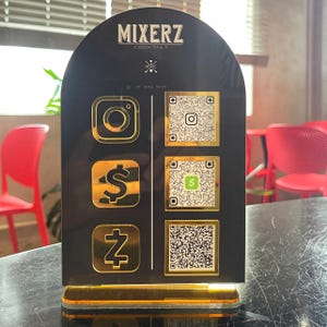 May include: A black arched acrylic sign with gold accents featuring the text MIXERZ COCKTAILS at the top. The sign displays three gold social media and payment icons on the left and three corresponding QR codes on the right, mounted on a base.