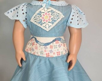 May include: A doll wearing a blue dress with white lace trim and a white embroidered belt. The doll has brown hair, green eyes, and is wearing black shoes.
